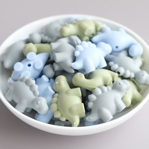 May include: A bowl filled with a variety of silicone dinosaur-shaped beads in shades of blue and green. The beads are perfect for sensory play and teething.