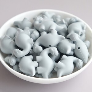 May include: A bowl filled with gray silicone dinosaur-shaped beads. The beads are all the same size and shape, and they are arranged in a random pattern.