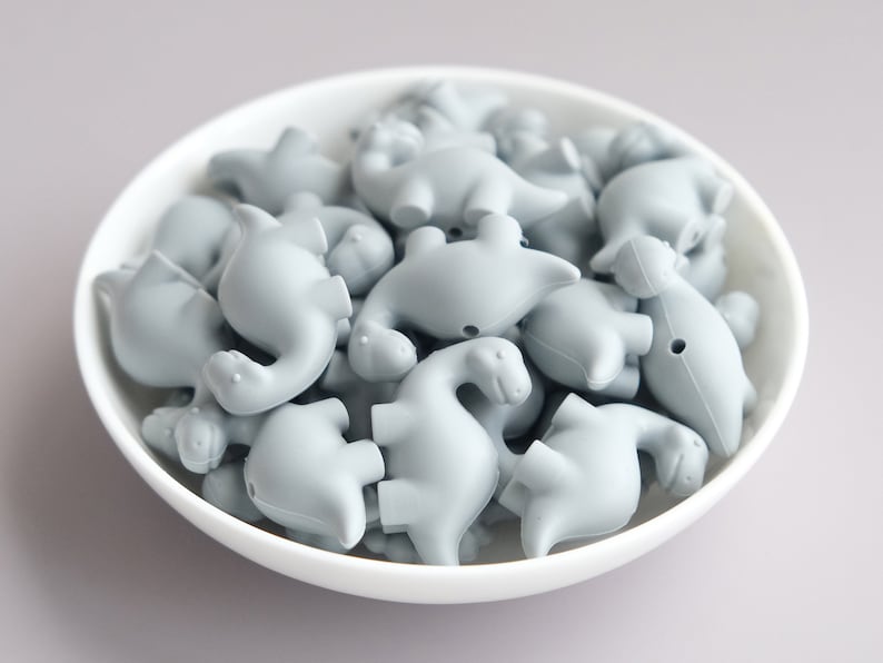 May include: A bowl filled with gray silicone dinosaur-shaped beads. The beads are all the same size and shape, and they are arranged in a random pattern.