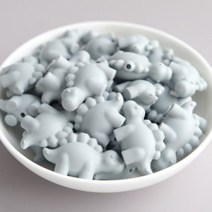 May include: A bowl filled with light gray silicone dinosaur-shaped beads. The beads are all the same size and shape, and they are arranged in a random pattern.