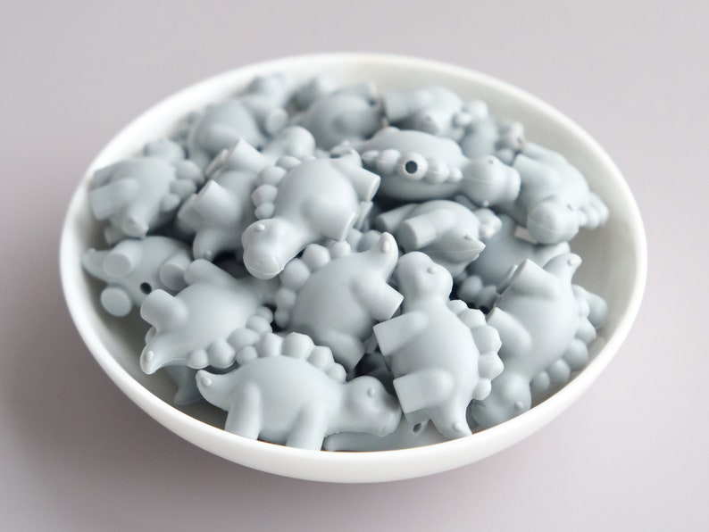 May include: A bowl filled with light gray silicone dinosaur-shaped beads. The beads are all the same size and shape, and they are arranged in a random pattern.
