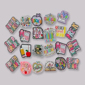 Op de afbeelding: Een verzameling kleurrijke charms met een thema voor leraren met glitteraccenten. Ontwerpen omvatten appels, boeken, potloden en zinnen als "Teach Love Inspire" en "Second Grade". Deze bedels zijn waarschijnlijk voor schoenen of accessoires.