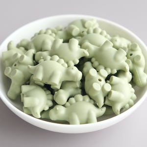 May include: A bowl filled with sage green silicone dinosaur-shaped beads. The beads are all the same size and shape, and they are arranged in a random pattern.