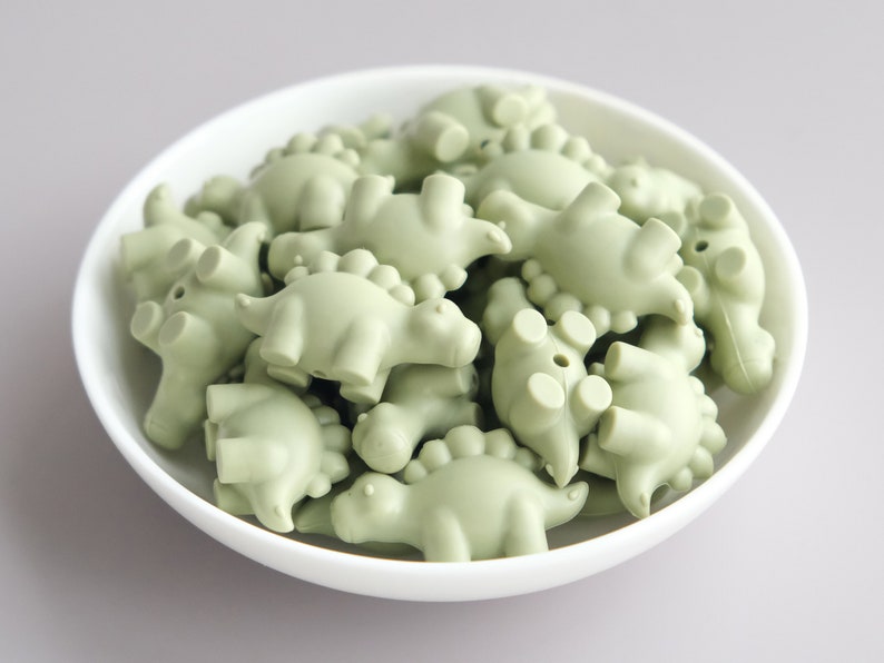 May include: A bowl filled with sage green silicone dinosaur-shaped beads. The beads are all the same size and shape, and they are arranged in a random pattern.