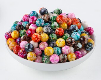 May include: A white bowl filled with a variety of colorful, marbled beads. The beads are round and come in shades of red, pink, yellow, green, blue, and black. These beads are ideal for jewelry making and crafting projects.