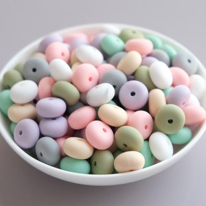Könnte beinhalten: Eine Schüssel gefüllt mit pastellfarbenen Silikonperlen. Die Perlen sind in den Farben Rosa, Grün, Lila und Grau. Sie sind alle rund und haben ein Loch in der Mitte.