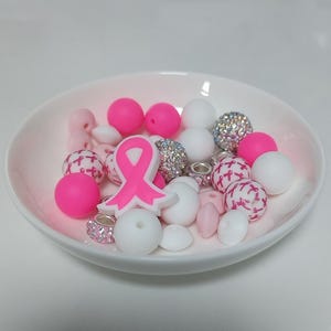 Könnte beinhalten: Eine weiße Schüssel gefüllt mit rosa und weißen Perlen. Die Perlen sind in verschiedenen Formen und Größen, darunter rund, flach und herzförmig. In der Mitte der Schüssel befindet sich ein rosa Schleifen-Anhänger.