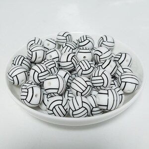 May include: A white bowl filled with white and black volleyball-themed beads. The beads are round and have a black outline of a volleyball.