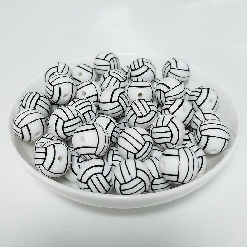 May include: A white bowl filled with white and black volleyball-themed beads. The beads are round and have a black outline of a volleyball.