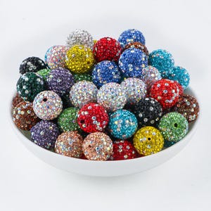 Pode incluir: Uma tigela branca cheia de uma variedade de contas coloridas cobertas de strass. As contas são em tons de vermelho, azul, verde, amarelo, roxo e castanho. Cada conta tem um buraco, sugerindo que são para fazer joias ou artesanato.