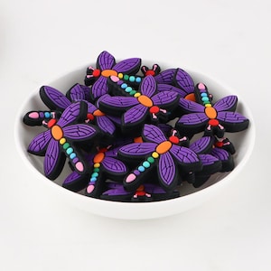 Op de afbeelding: Een kom vol met paarse libellenbedels met kleurrijke vleugels. De bedels zijn gemaakt van een zacht, flexibel materiaal en perfect om een vleugje whimsy toe te voegen aan elk project.