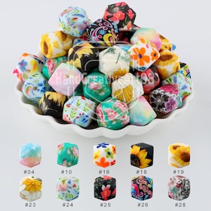 Könnte beinhalten: Eine Sammlung von 28 bunten, quadratischen Silikonperlen mit verschiedenen floralen und geometrischen Mustern. Die Perlen sind in einer weißen Schüssel angeordnet. Jede Perle hat ein Loch in der Mitte zum Auf fädeln.