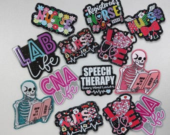 May include: A collection of colorful, glittery pins with medical and nursing themes. Designs include the words "Nurse," "Lab Life," "CNA Life," and "Speech Therapy." Some pins feature stethoscopes, hearts, and a skeleton reading a book.