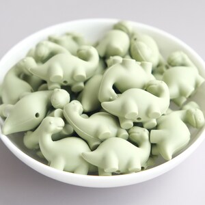 May include: A bowl filled with pale green silicone dinosaur-shaped beads. The beads are all the same size and shape, and they are arranged in a random pattern.