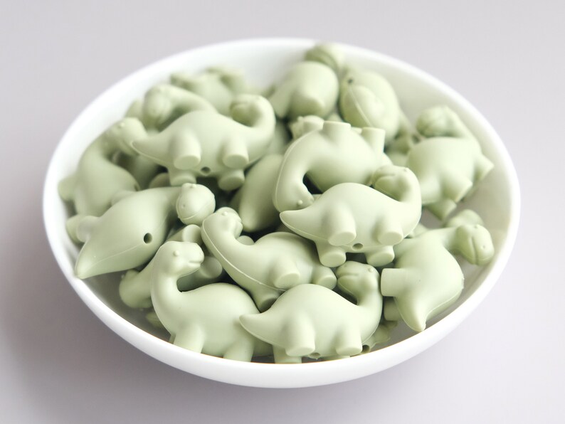 May include: A bowl filled with pale green silicone dinosaur-shaped beads. The beads are all the same size and shape, and they are arranged in a random pattern.