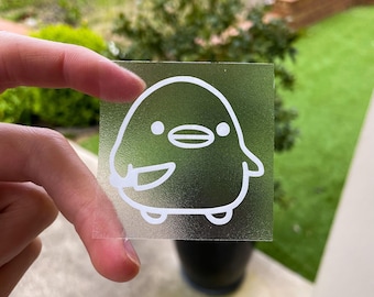 May include: A clear sticker with a white outline of a cartoon penguin holding a knife. The penguin has a small beak and two small eyes.