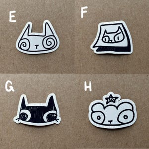 May include: Four black and white stickers featuring different animal designs. The stickers are on a brown surface. The designs include a cat with swirls, a cat wearing a hat, a cat with a black and white face, and a panda with a star.