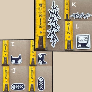 May include: A set of nine black and white stickers featuring various designs, including a cat, a fishbone, and a stylized tree. The stickers are arranged on a brown surface with a yellow measuring tape.