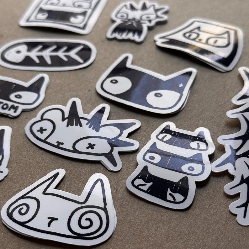 May include: A collection of black and white cat stickers with various designs, including a cat with a bone, a cat with a skull, and a cat with a spiral design. The stickers are on a light gray surface.