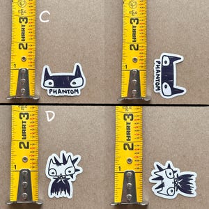 May include: Four black and white stickers featuring cartoon cats. Two of the stickers feature a cat with a mohawk and the other two feature a cat with a bat-like mask. One of the cat stickers with a bat-like mask has the word "PHANTOM" written below it.