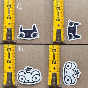 May include: Four black and white stickers with cartoon animal designs. The stickers are on a brown surface with a yellow measuring tape on either side. The stickers are labeled G and H. The stickers feature a cat, a cat with a star, a bear with a star, and a frog.