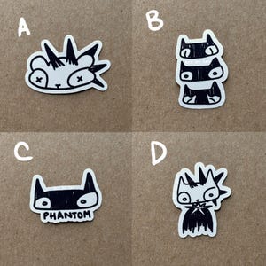 May include: Four black and white cat stickers on a brown background. The stickers feature different cat designs, including a cat with a mohawk, a stack of three cats, a cat with the word "PHANTOM" underneath, and a cat with a mohawk and a cape.