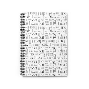 May include: A white notebook with a spiral binding features a repeating pattern of airport codes in black text. The codes include ATL, ORD, JFK, LAX, and more.