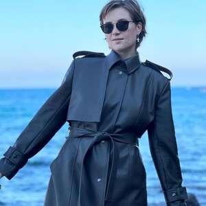 May include: A woman wearing a dark gray leather trench coat with a belt. She is standing in front of a blue body of water.