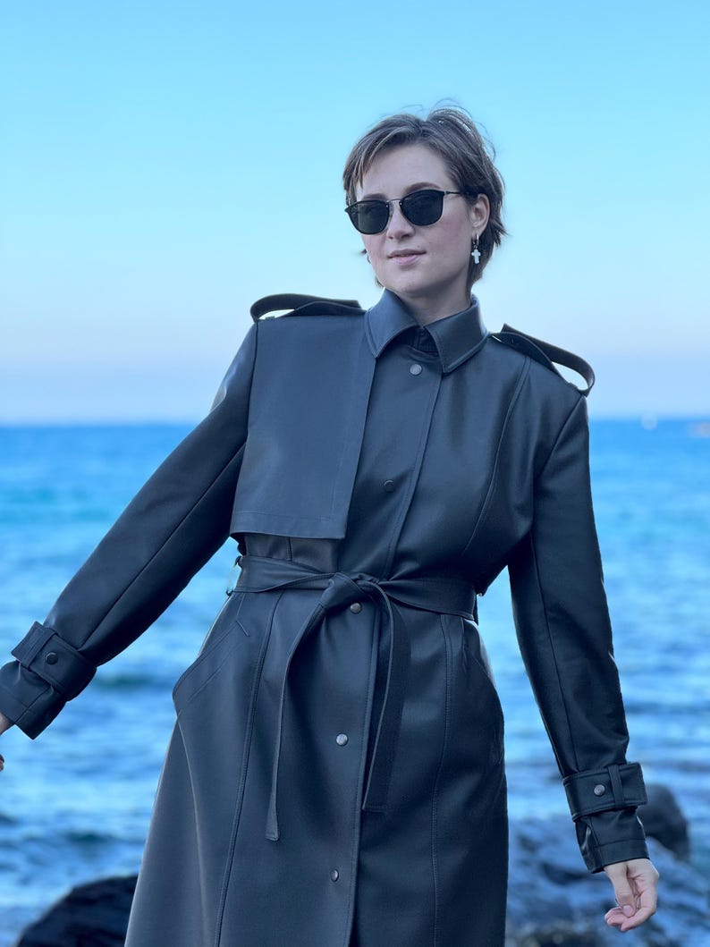 May include: A woman wearing a dark gray leather trench coat with a belt. She is standing in front of a blue body of water.