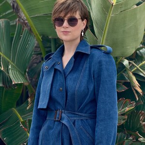 May include: A woman wearing a blue denim trench coat with a brown belt. She is wearing sunglasses and has short brown hair.