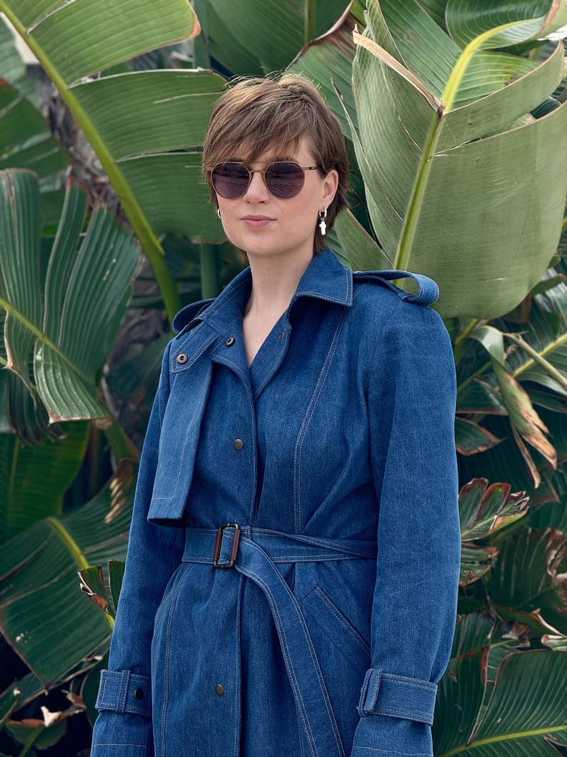 May include: A woman wearing a blue denim trench coat with a brown belt. She is wearing sunglasses and has short brown hair.