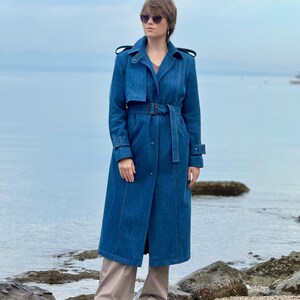 May include: A woman wearing a long, blue denim trench coat with a belt, standing on a rocky beach. She is wearing sunglasses and has short brown hair.