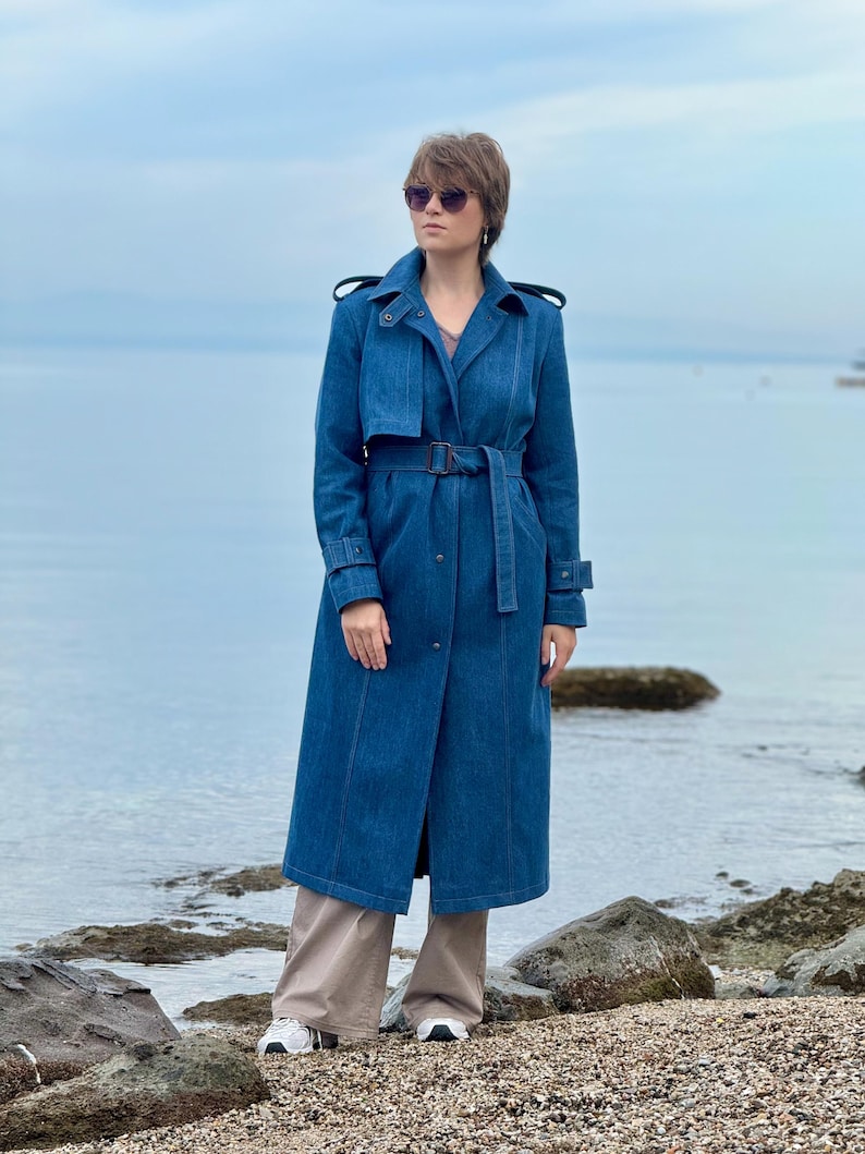 May include: A woman wearing a long, blue denim trench coat with a belt, standing on a rocky beach. She is wearing sunglasses and has short brown hair.