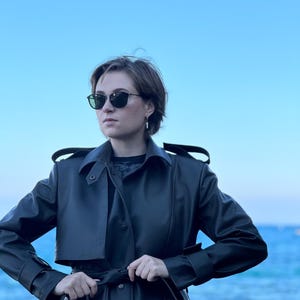 May include: A woman wearing a black leather trench coat with a belt, standing in front of a blue body of water. She is wearing sunglasses and has short brown hair.
