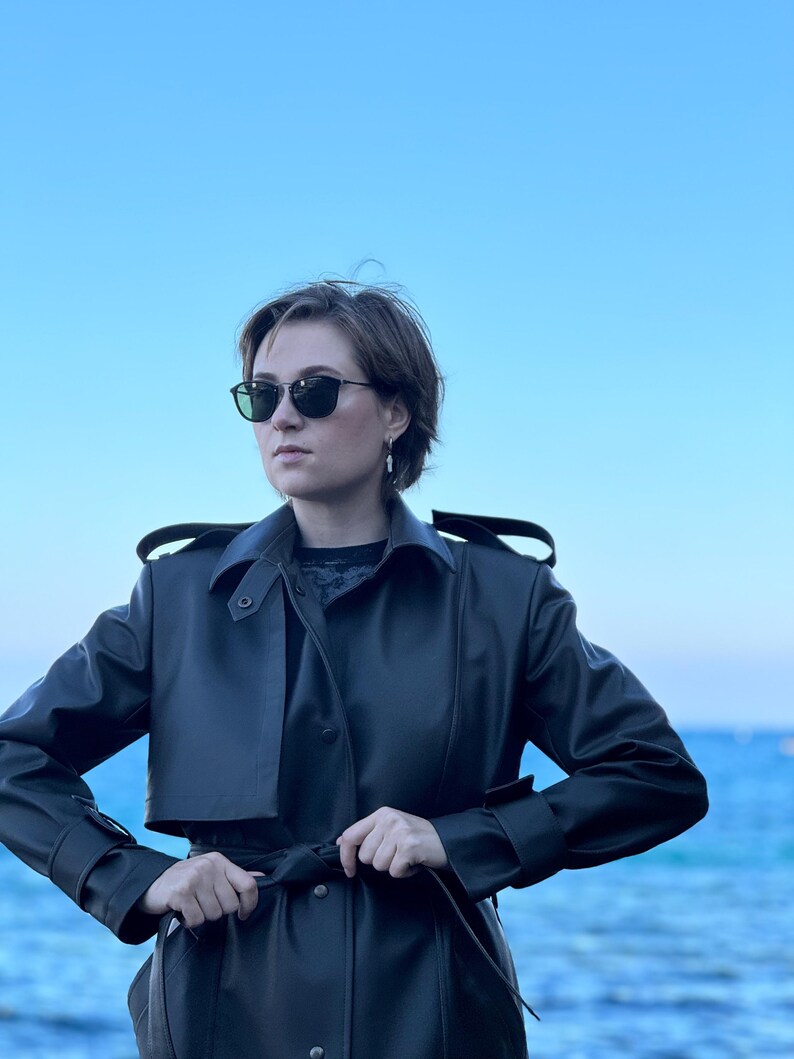 May include: A woman wearing a black leather trench coat with a belt, standing in front of a blue body of water. She is wearing sunglasses and has short brown hair.