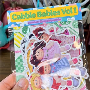 May include: A pack of stickers featuring Cabbage Patch Kids-style dolls in various outfits. The stickers are in a clear plastic sleeve with the text "Cabbie Babies Vol 1" and the Etsy shop name. The dolls have different hair colors and wear dresses.