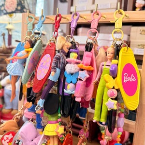 May include: A collection of keychains featuring Barbie dolls and small plush toys. The keychains have various designs, including Barbie-themed tags, colorful beads, and miniature figures. The keychains are displayed on a wooden rack.
