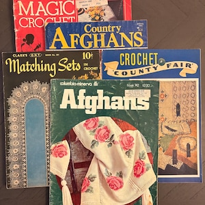 May include: A selection of vintage crochet pattern books. Titles include "Magic Crochet," "Country Afghans," "Matching Sets," and "Crochet County Fair." The "Afghans" book features a floral design. The books are in various colours.