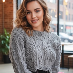 Könnte beinhalten: Ein grauer, gecroppter Zopfstrickpullover mit langen Ärmeln. Der Pullover hat ein strukturiertes Muster und wird mit einem karierten Rock kombiniert. Das Bild enthält ein Shop-Logo in der unteren rechten Ecke.