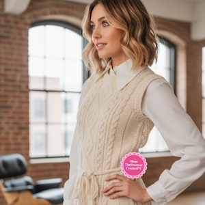 Op de afbeelding: Crèmekleurig kabelgebreid vest met een bijpassende strikriem en zijsplitten. Het vest wordt gedragen over een wit overhemd met kraag en een donkerblauwe jeans. De afbeelding bevat de tekst "Shop: TheSweetest CrochetPT".