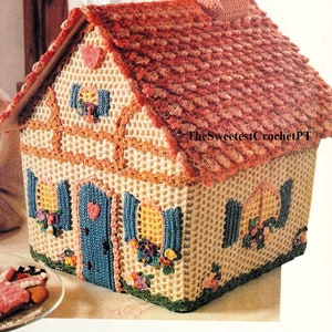 Puede incluir: Cubierta de caja de pañuelos de tejido de punto en forma de casa con un techo rosa, una puerta azul y ventanas amarillas. La casa está decorada con flores y corazones tejidos.