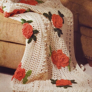 May include: A crocheted throw blanket with a white background and orange roses. The blanket is draped over a brown couch.
