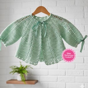 Könnte beinhalten: Ein mintgrünes Häkeltop mit langen Ärmeln und einem gewellten Saum. Das Top hat einen runden Ausschnitt mit einer Schleife und Bändern, die an den Manschetten gebunden sind. Das Top hängt an einem Holzbügel.