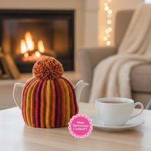Puede incluir: Una tetera blanca con una funda de punto en tonos rojos, naranjas y amarillos, rematada con un pompón. Una taza y un platillo blancos están cerca. La funda tiene rayas verticales. El fondo incluye una chimenea y una silla acogedora.