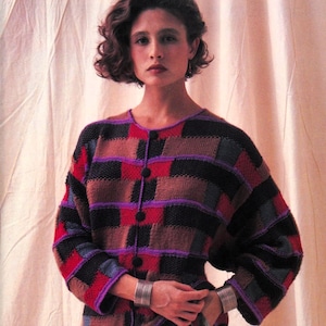 Op de afbeelding: Een vrouw draagt een veelkleurige gebreide trui met een knoopsluiting. De trui heeft een geruit patroon in tinten bruin, rood, paars en zwart. De vrouw draagt een gestreepte rok en zilveren armbanden.