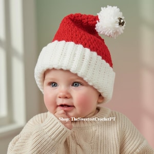 Puede incluir: Un gorro de Papá Noel rojo y blanco tejido a ganchillo, con un pompón blanco y una pequeña campana plateada. El gorro lo lleva un bebé que lleva un jersey color crema. El texto "Shop: TheSweetestCrochetPT" es visible.