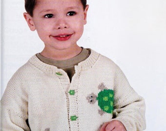 May include: A cream-colored knit cardigan featuring a crew neckline, long sleeves, and a button-down front. The sweater is decorated with green turtle motifs and small green turtle-shaped buttons. The text Shop: TheSweetestCrochetPT is visible.