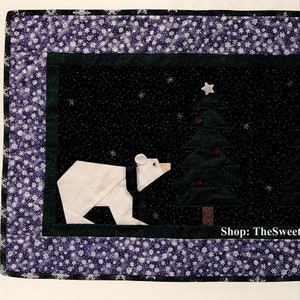 May include: A quilted Christmas table runner featuring a dark green and black scene with three white polar bears, a Christmas tree, and a star. The border is a blue fabric with white snowflakes. The text "Shop: TheSweetestCrochetPT" is visible.
