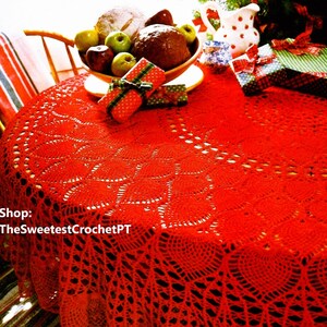 May include: Red crocheted table cloth with a delicate lace pattern. The tablecloth is laid on a table with a bowl of fruit, wrapped gifts, and a pitcher on top.