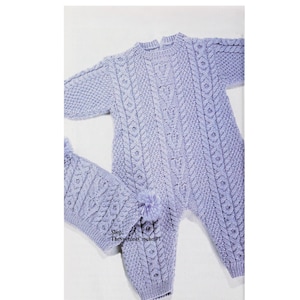 May include: A light purple knitted baby onesie with a cable knit pattern and pom poms on the feet. The onesie is laid out on a white surface. Shop: TheSweetestCrochetPT